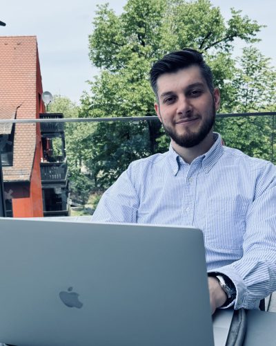 Ein Geschäftsführer der Marketingagentur OMNI2 sitzt vor einem Laptop und lächelt in die Kamera. Er trägt ein gestreiftes Hemd, eine Uhr und hat einen vollen Bart.