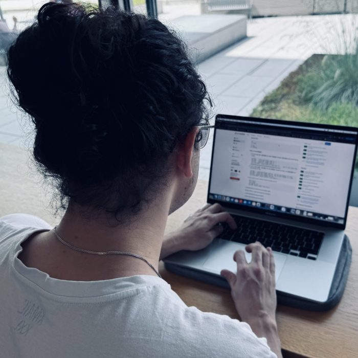 Ein Foto zeigt einen Mitarbeiter von OMNI2 von hinten, während er am MacBook arbeitet. Er trägt ein weißes Shirt und hat seine Haare zu einem Zopf gebunden.